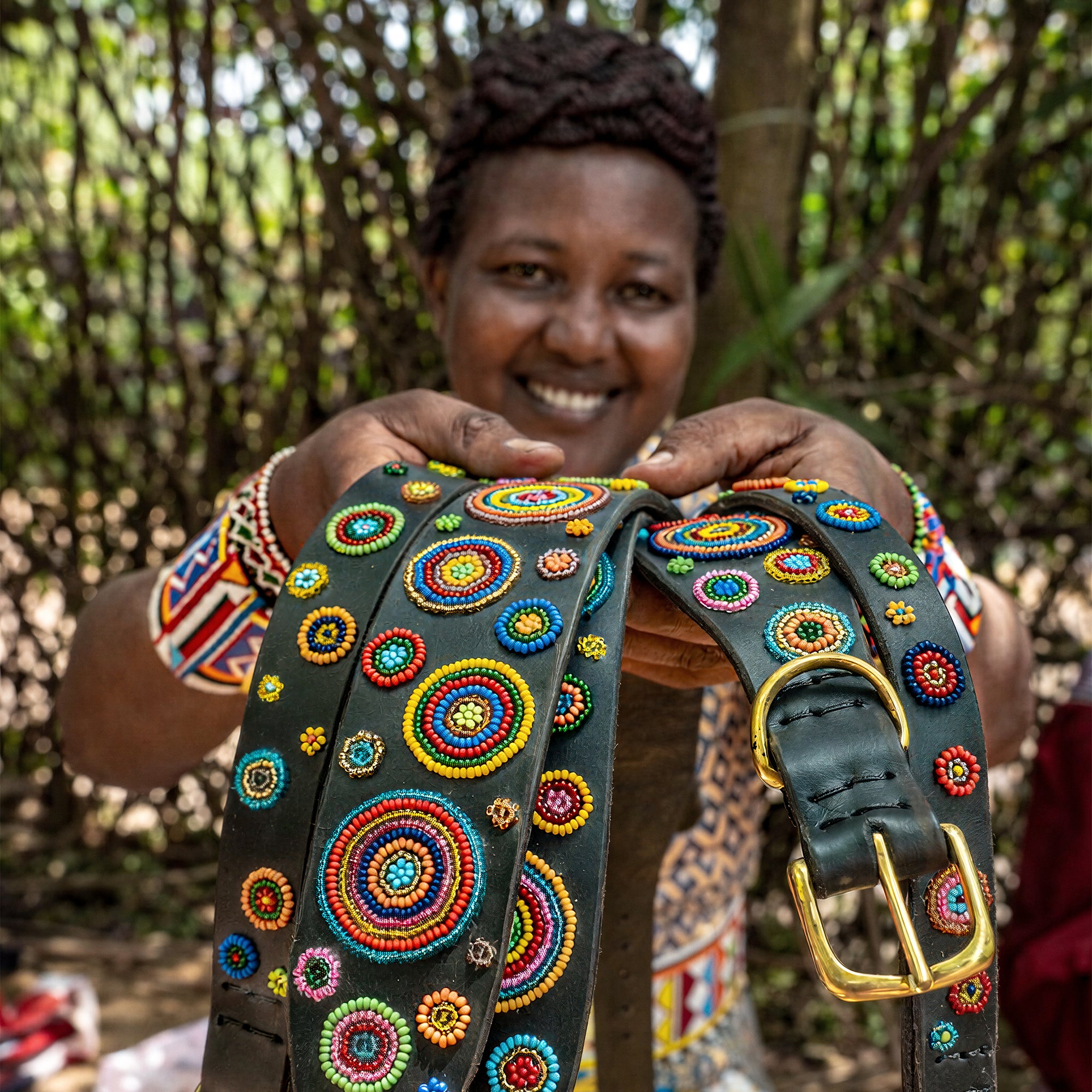 a Woman from the Maasa Mara in traditional attire sitting outdoors showing her handmade colorful dog collars from The Maasai Collection 