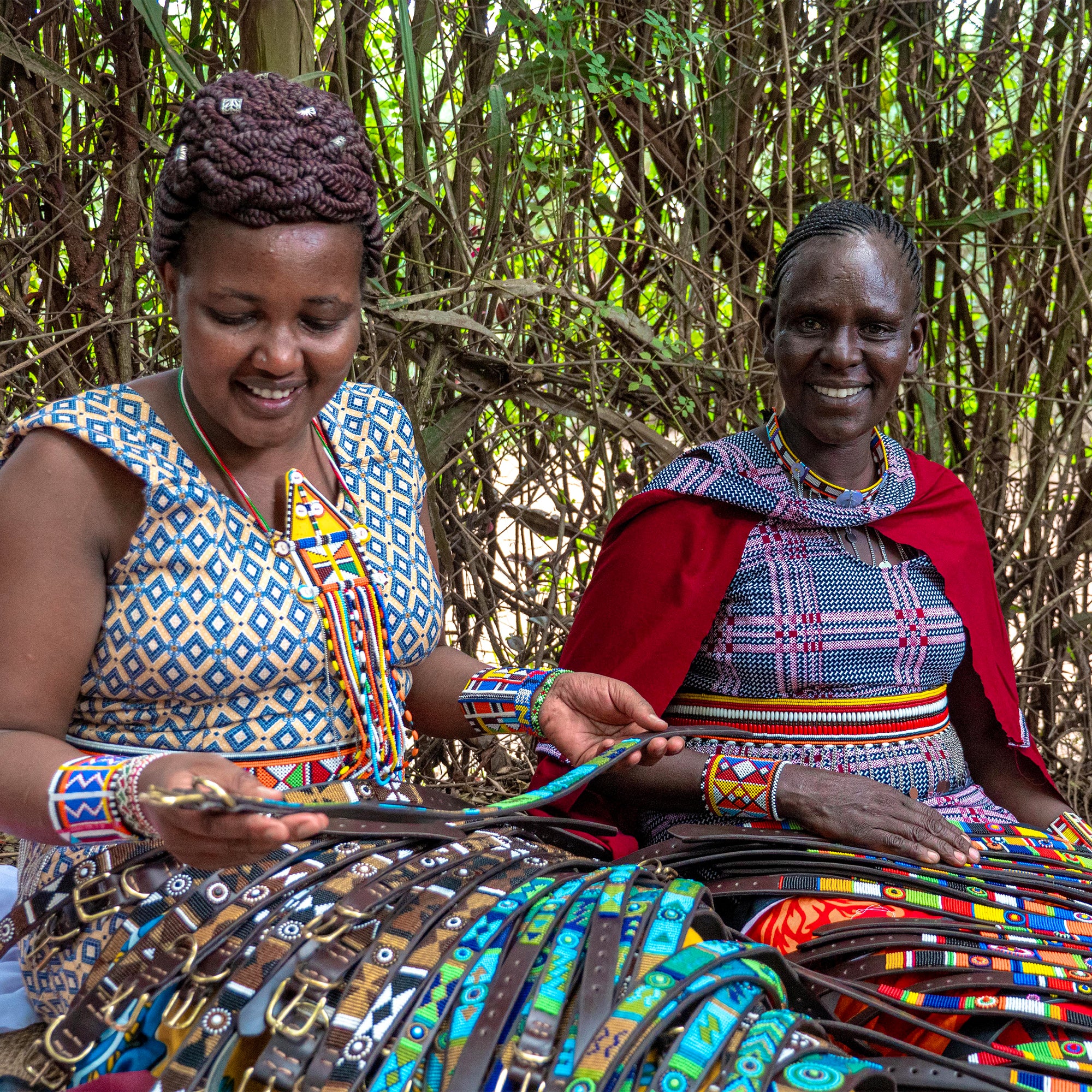 Two women from the Maasa Mara in traditional attire sitting outdoors with colorful handmade dog collars from The Maasai Collection 