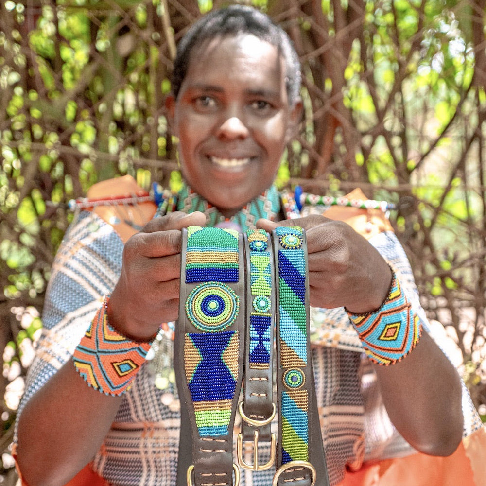 Maasai woman in traditional clothing showcasing her handmade dog collars for The Maasai Collection featuring the blue green and gold Peacock design 